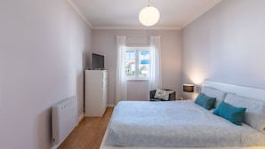 Ett sovrum med en säng, en tv på ett skåp, ett fönster med gardiner, en lampa och ett element.