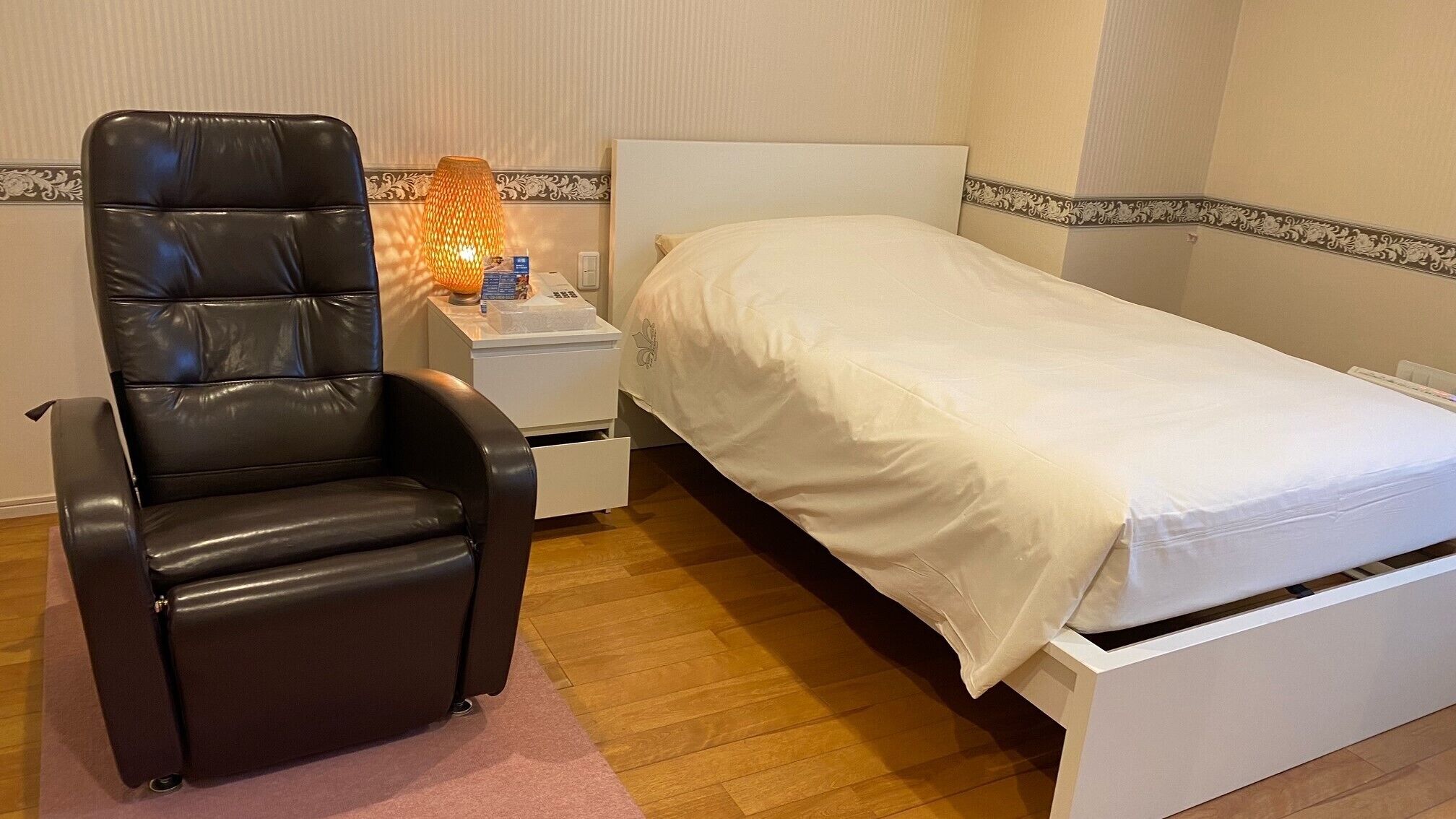 A small, single-bed room with a black leather chair, a bedside table with a lamp, and a white bedspread.