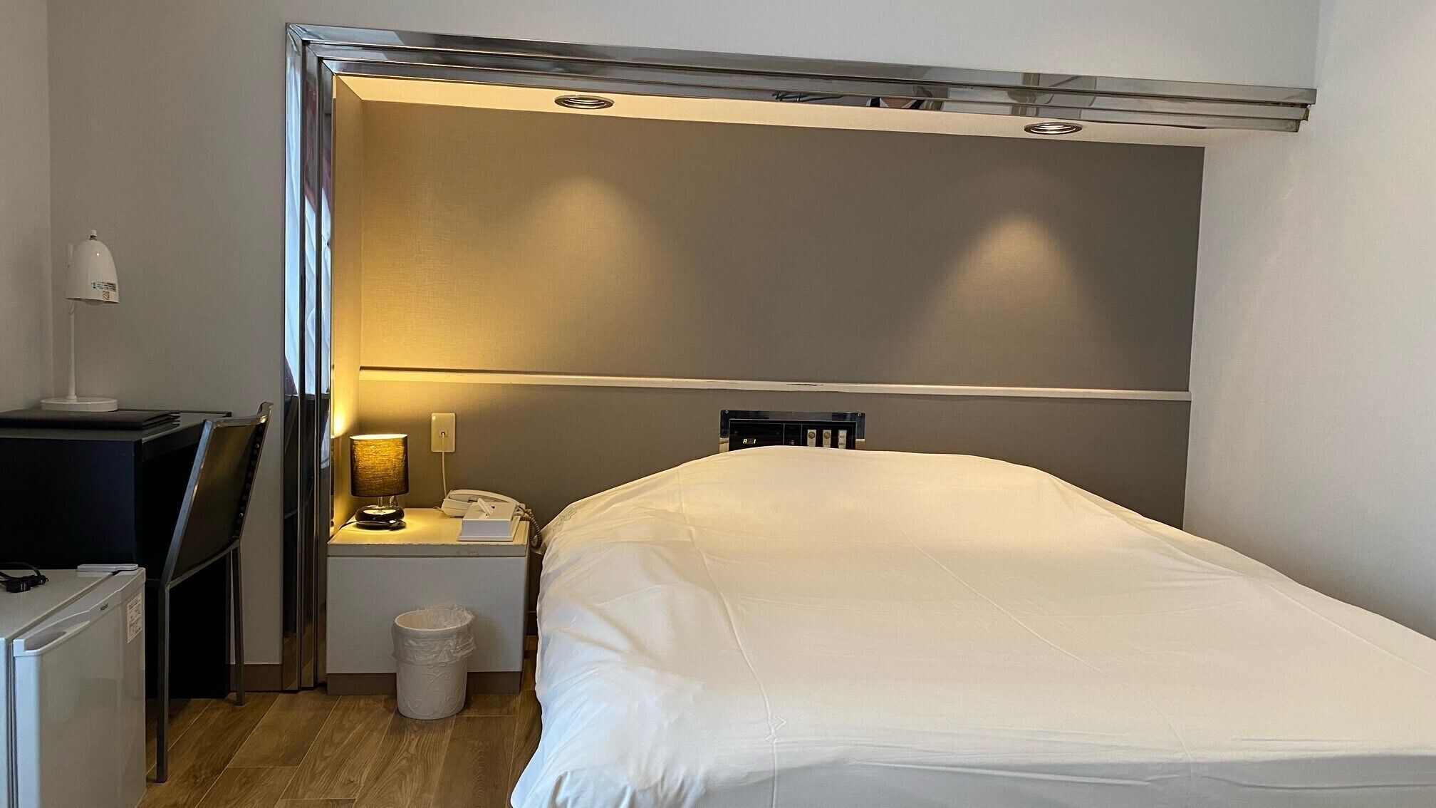 A compact hotel room with a bed, a desk with a lamp, a chair, and a small trash bin.