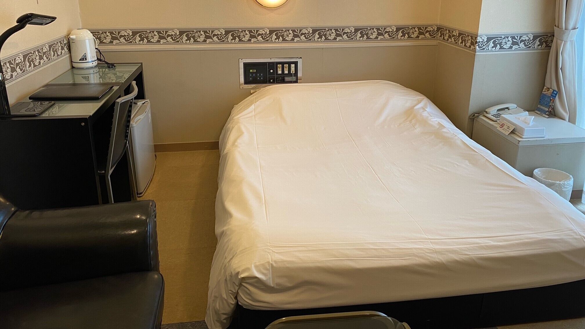 A hotel room with a bed, a desk with a lamp, a telephone, and a trash bin.