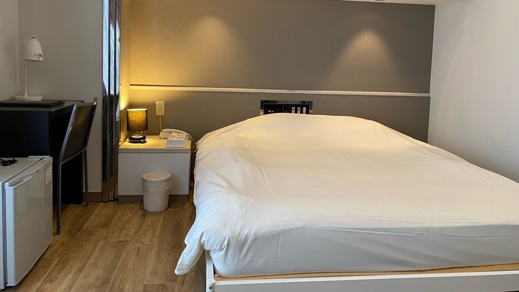 A hotel room with a bed, a desk, a lamp, a trash bin, and a wall-mounted phone.