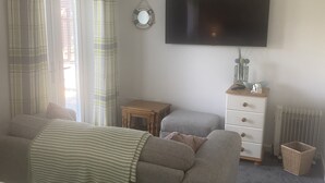 A bedroom with a bed, a grey sofa, a small wooden table, a TV mounted on the wall, and a radiator. - The Batch, 5* Self Catering Bungalow, St Merryn Padstow (St Merryn)