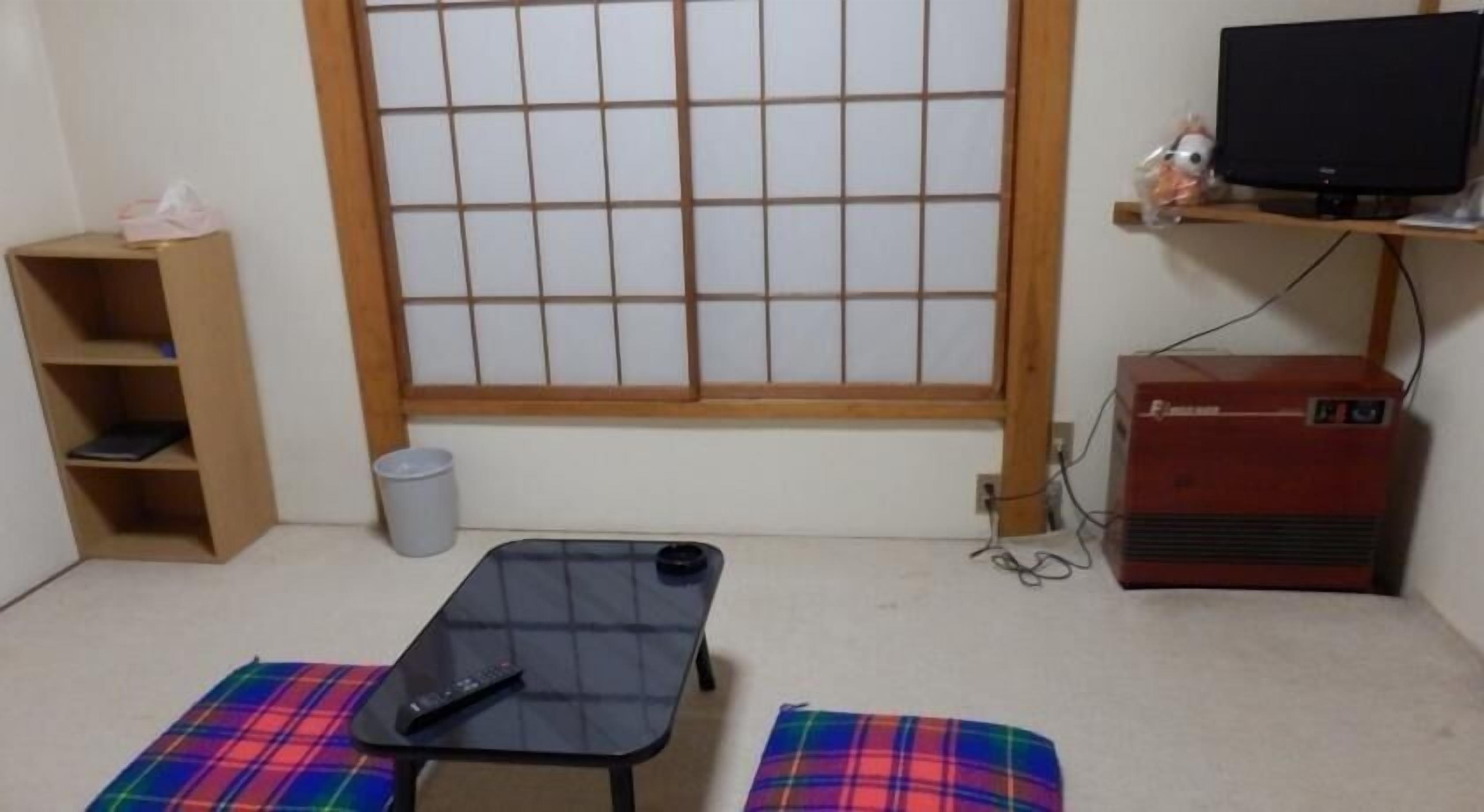 A room with a futon bed, a television on a shelf, a small heater, and a window with a grid pattern.