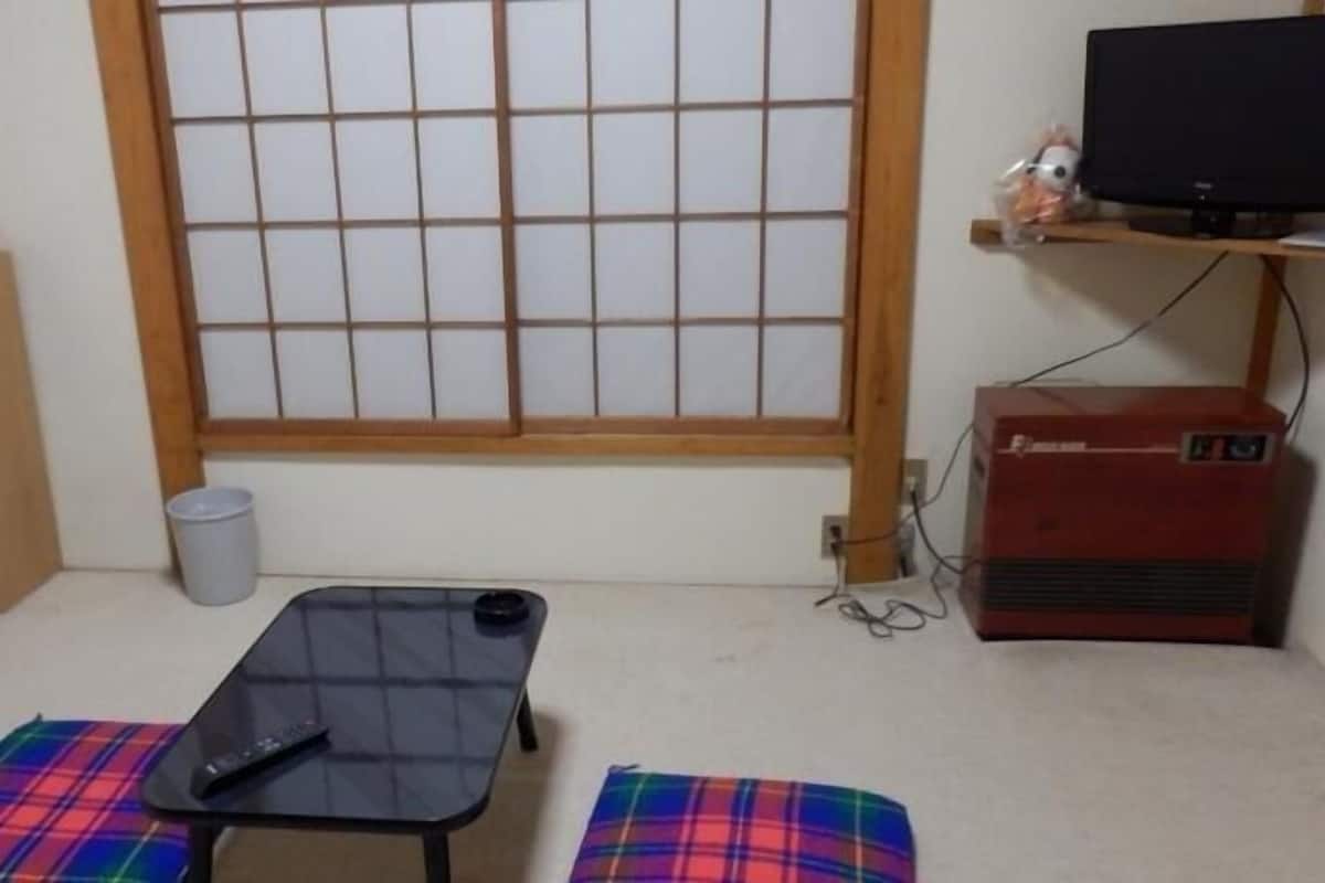 Ein Zimmer mit Futonbett, Fernseher auf einem Regal, kleinem Heizgerät und Fenster mit Gittermuster.