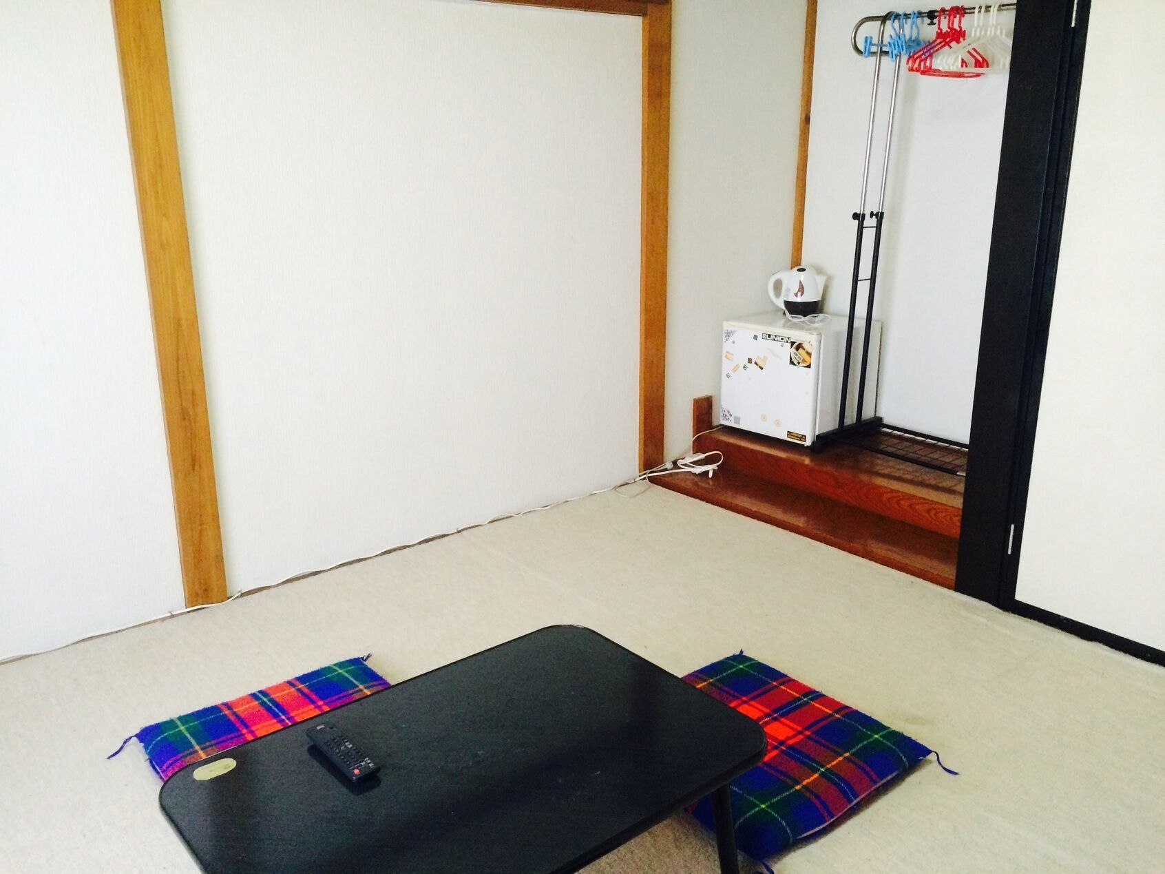 A room with a bed, a small table, a remote control, a kettle, and a vacuum cleaner.