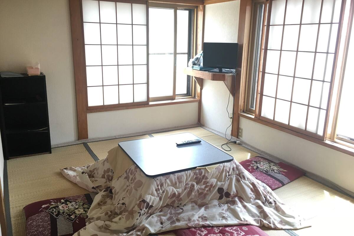 Ein traditioneller japanischer Raum mit Tatami-Böden, einem Futonbett, einem kleinen schwarzen Schrank und einem Fernseher auf einem Holzboden.