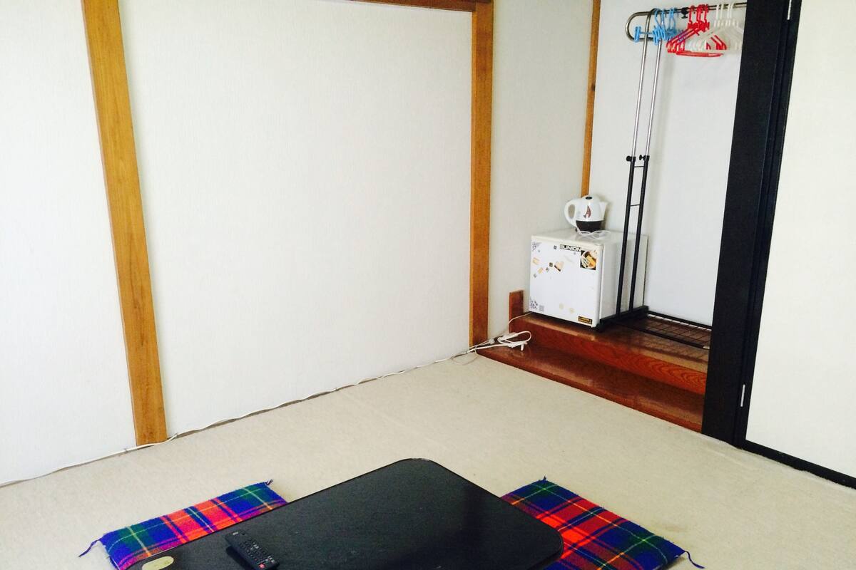 Ein Zimmer mit einem schwarzen Futon, einer Fernbedienung, einer farbenfrohen Karo-Decke, einem weißen Heizkörper und einem weißen Gerät mit Aufkleber.