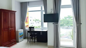 Quarto de hotel com guarda-roupa de madeira, pequeno frigorífico, secretária com cadeira, televisão na parede e grandes janelas com cortinas.