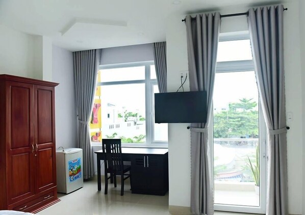 A hotel room with a wooden wardrobe, a small refrigerator, a desk with a chair, a TV mounted on the wall, and large windows with curtains.