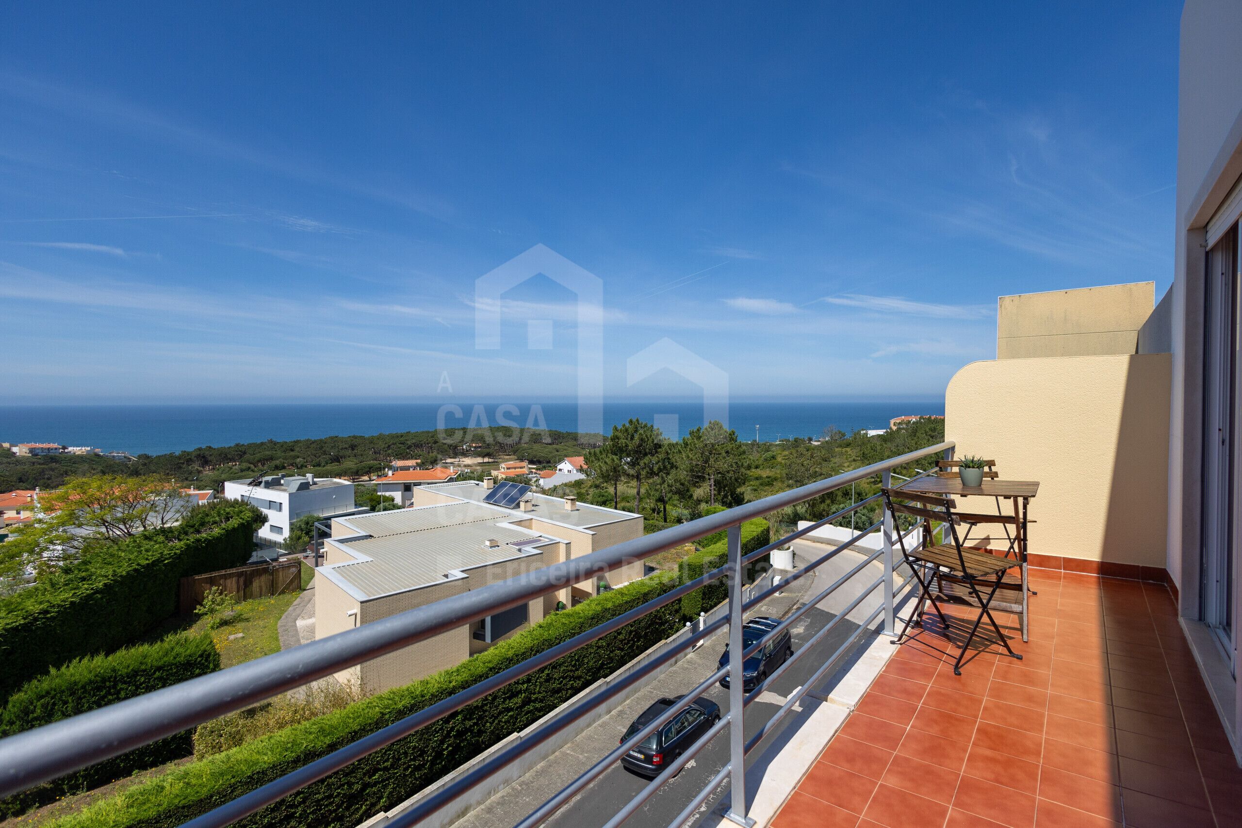 A balcony with a view of the sea, a table with chairs, and a car parked on the street.