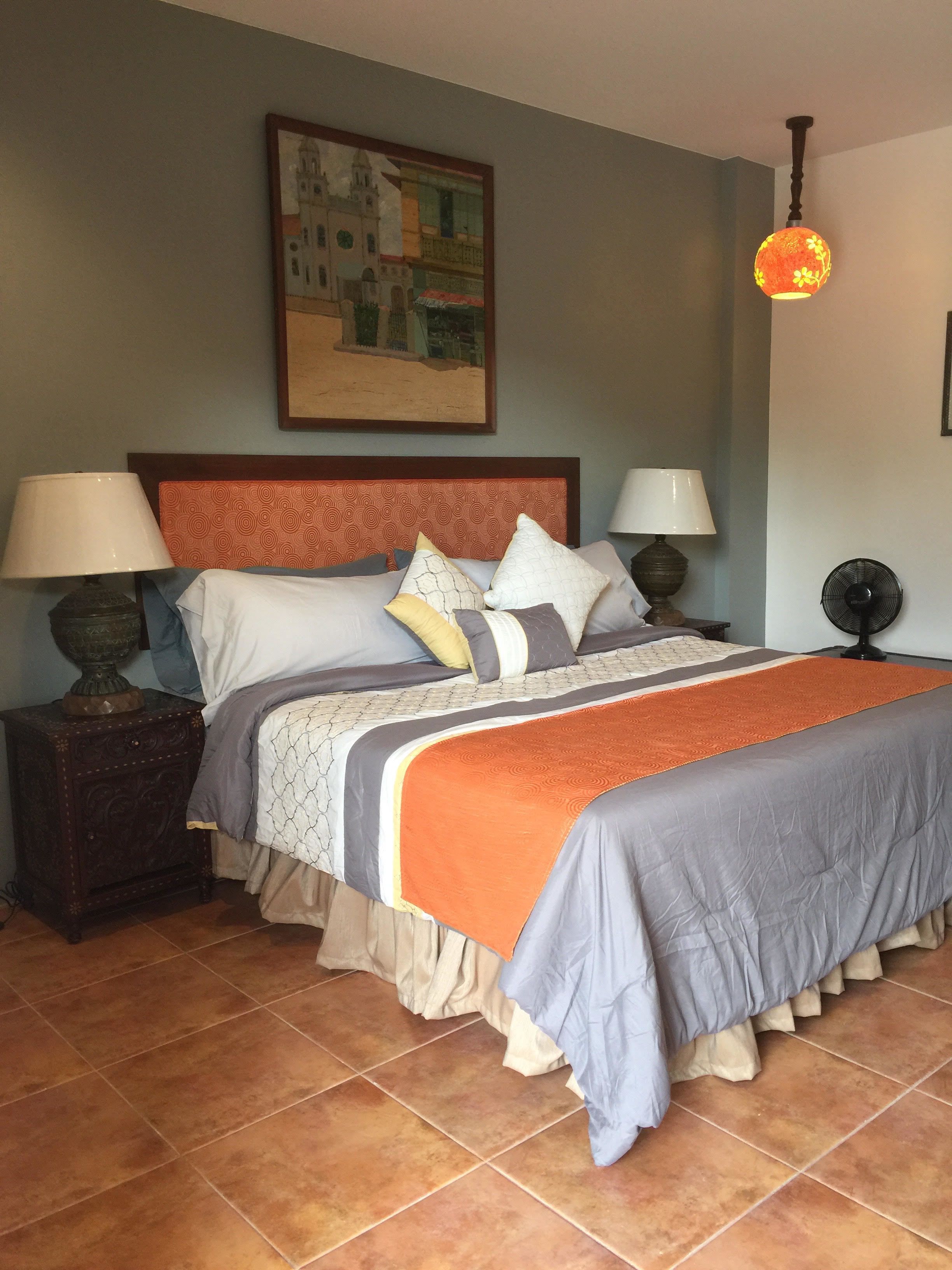 Uma cama bem arrumada com cabeceira estampada, duas mesas de cabeceira com abajures, um ventilador fixado na parede e um quadro sobre a cama.