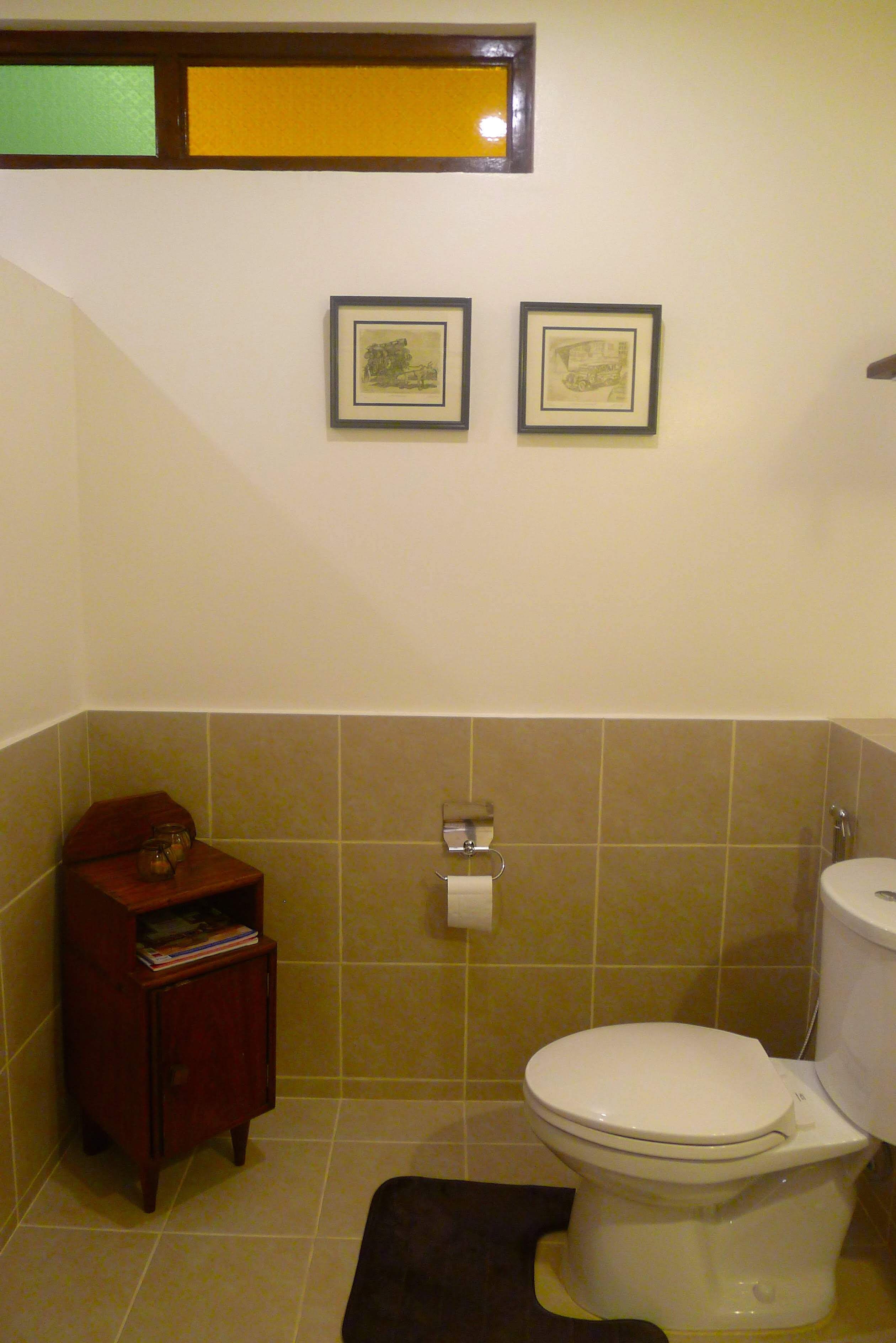 A bathroom with a toilet, a small wooden cabinet with a book, and two framed pictures on the wall.