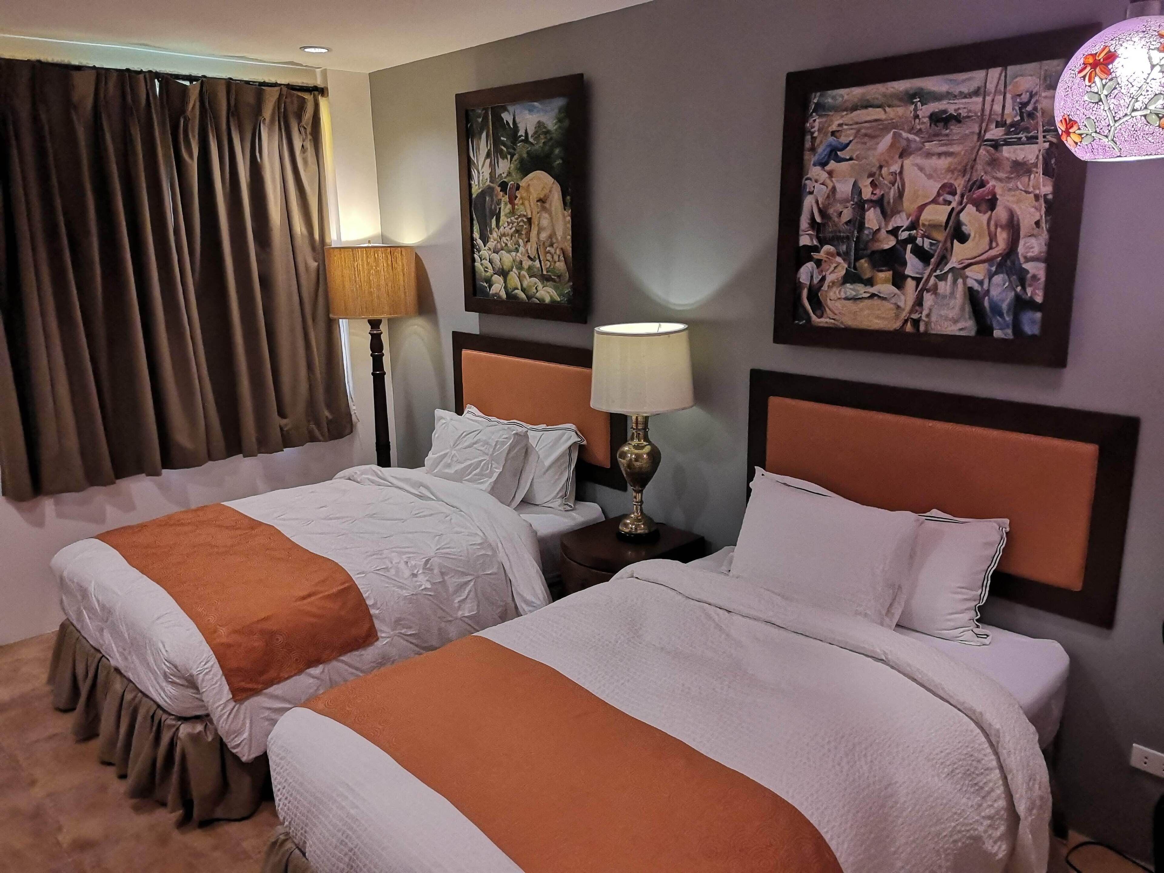 Habitación de hotel con dos camas, ventana con cortinas, una lámpara y obras de arte en las paredes.