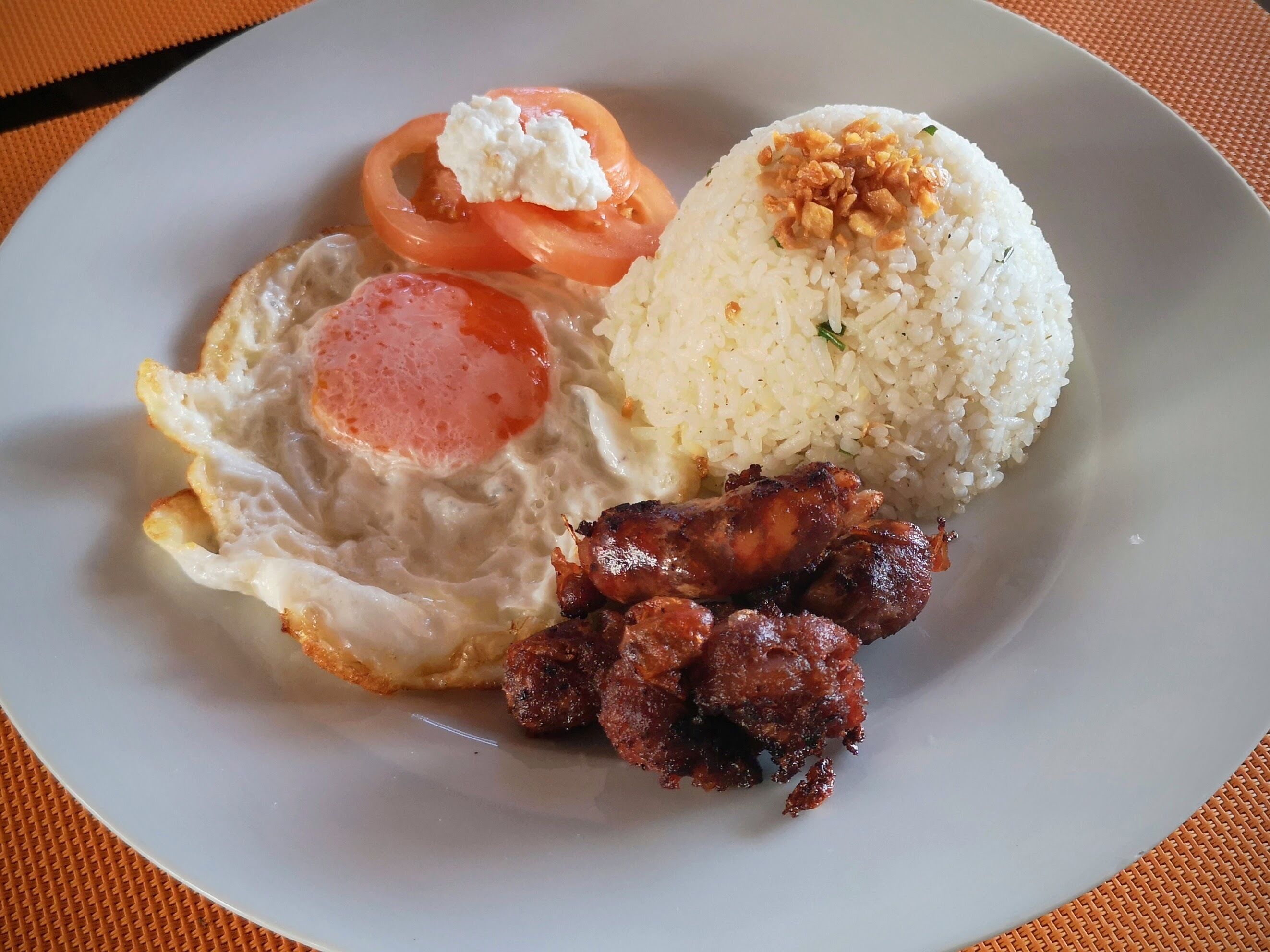 A plate with fried egg, sausage, rice, and a slice of tomato.