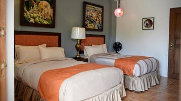 Una habitación de hotel con dos camas, una mesita de noche, una lámpara, un ventilador y obras de arte en las paredes.