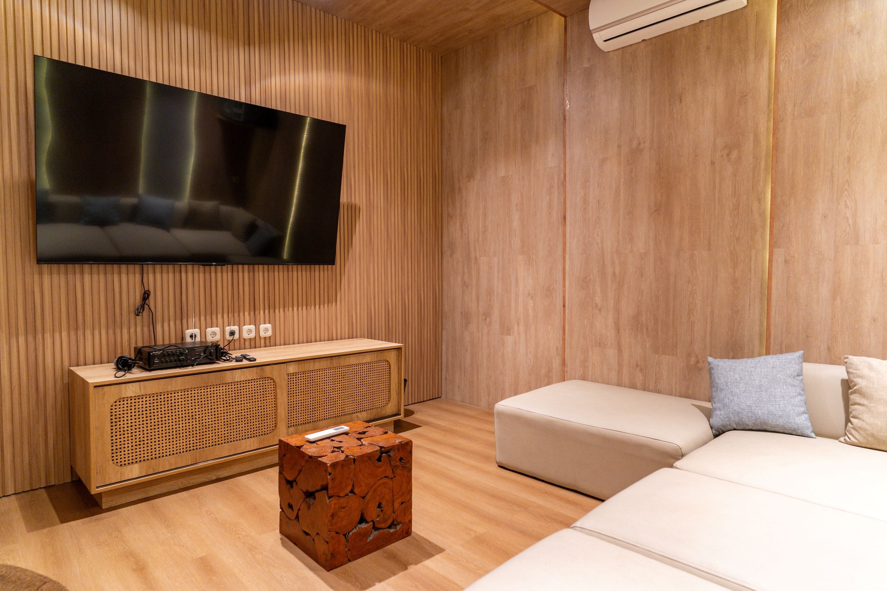 Una sala de estar moderna con un televisor de pantalla plana montado en la pared, un mueble bajo de madera con dispositivos electrónicos, una mesa de centro de madera original y un sofá beige con cojines.