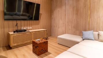 Una sala de estar moderna con un televisor de pantalla plana montado en la pared, un mueble bajo de madera con dispositivos electrónicos, una mesa de centro de madera original y un sofá beige con cojines.