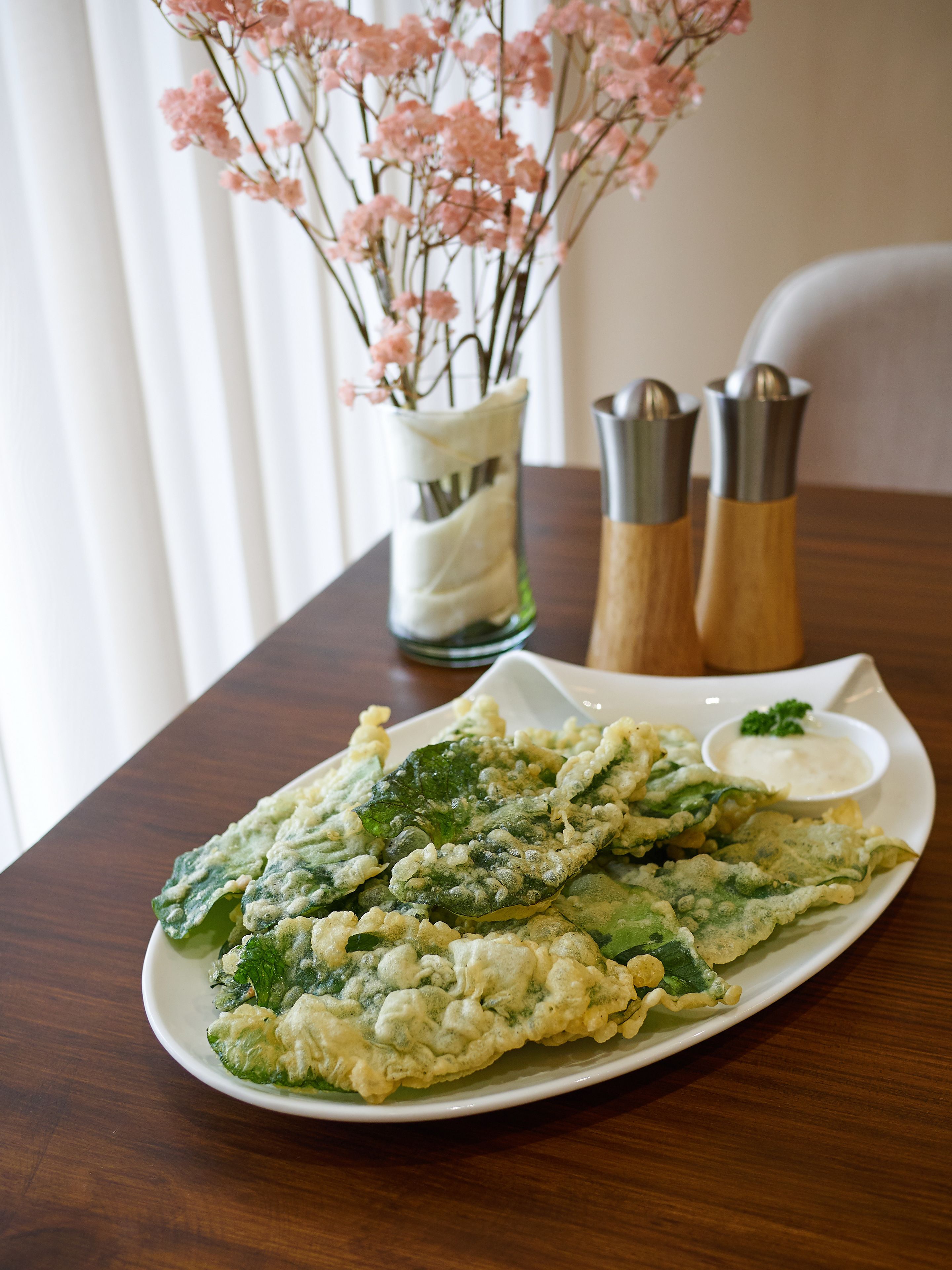 A plate of tempura with a dipping sauce, a small bowl of sauce, and a vase with pink flowers on a wooden table.