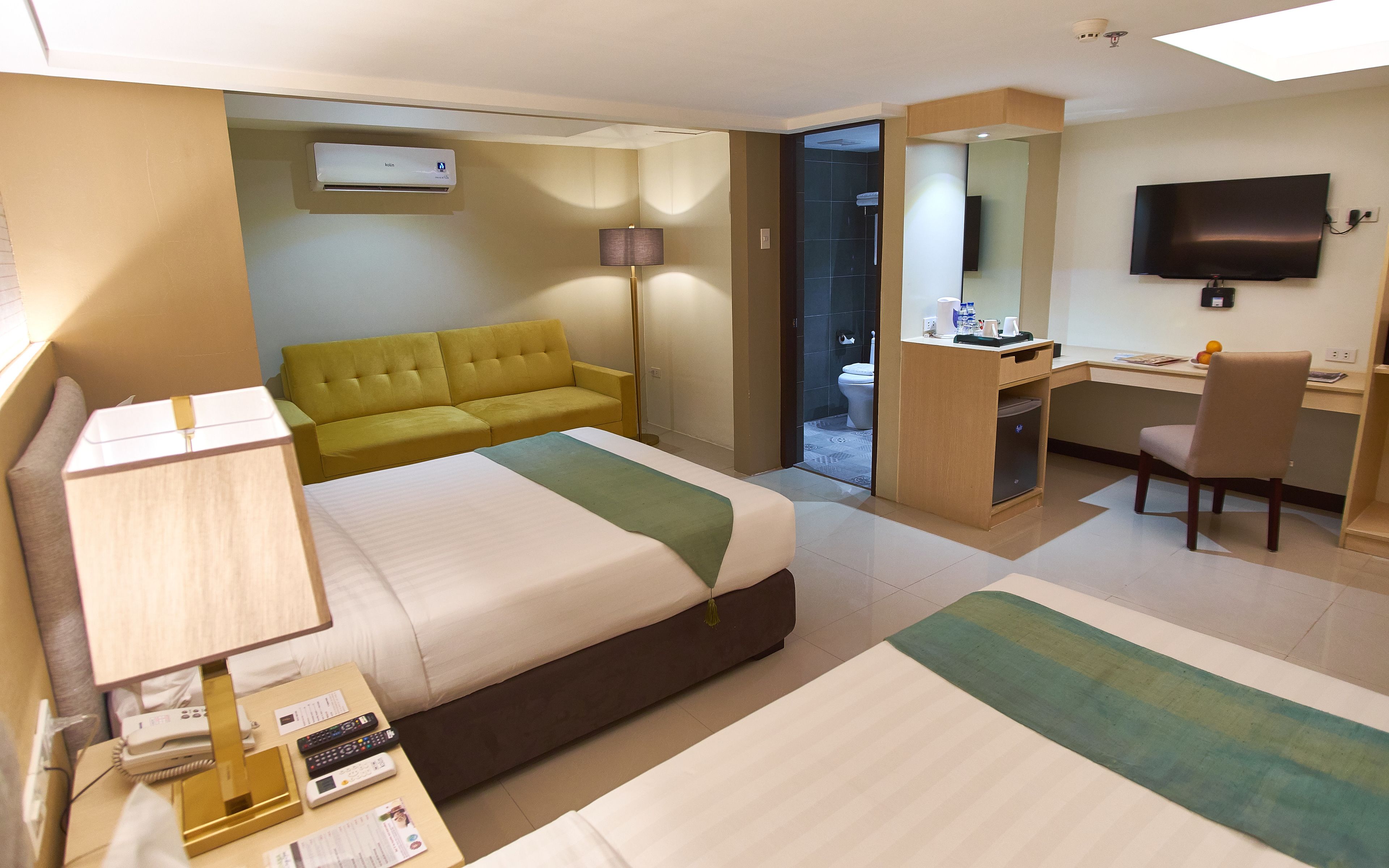 A hotel room with a bed, a desk, a chair, a sofa, a TV, and a bathroom visible through an open door.