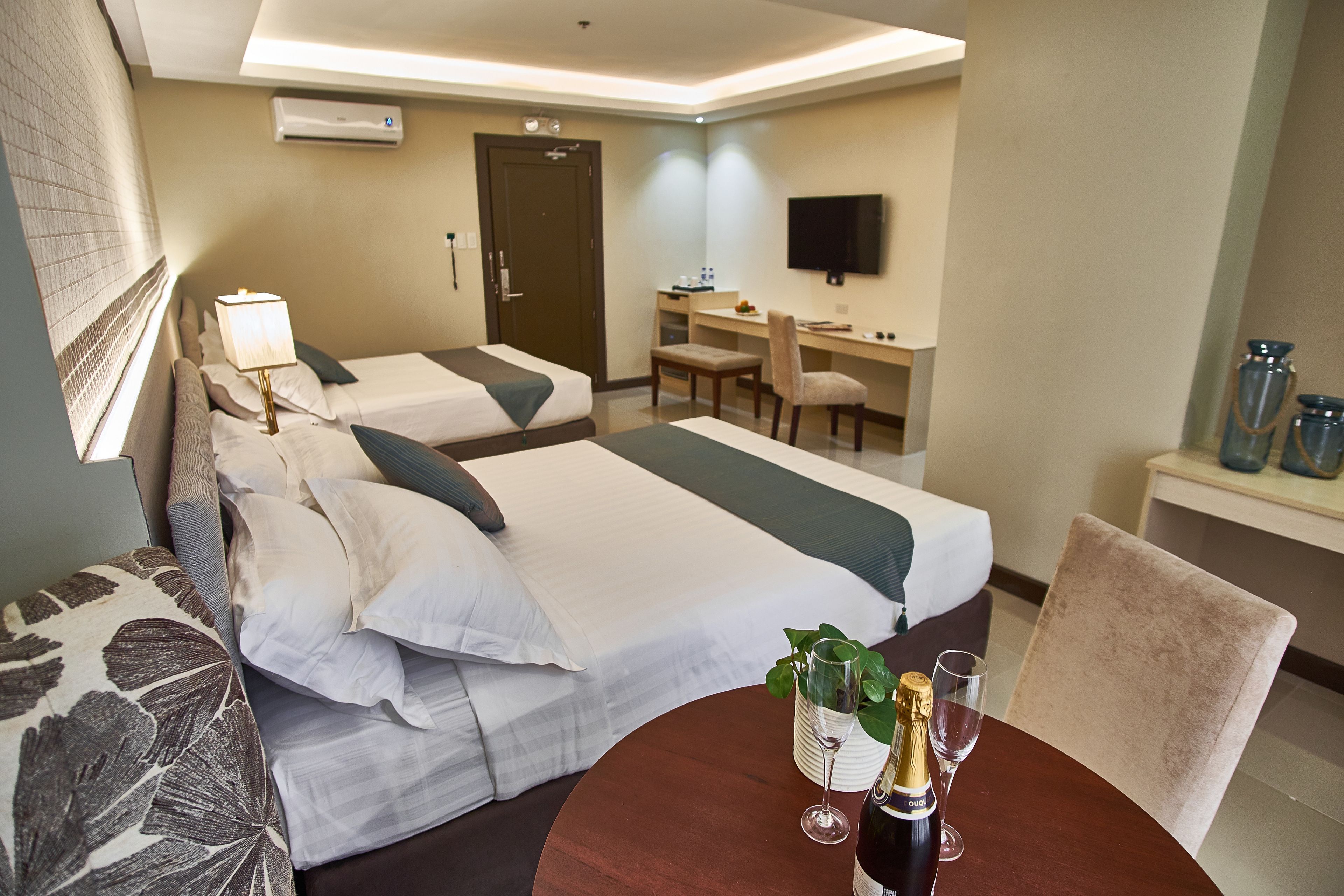 A hotel room with two beds, a desk, a chair, a TV, and a table with a bottle and glass.
