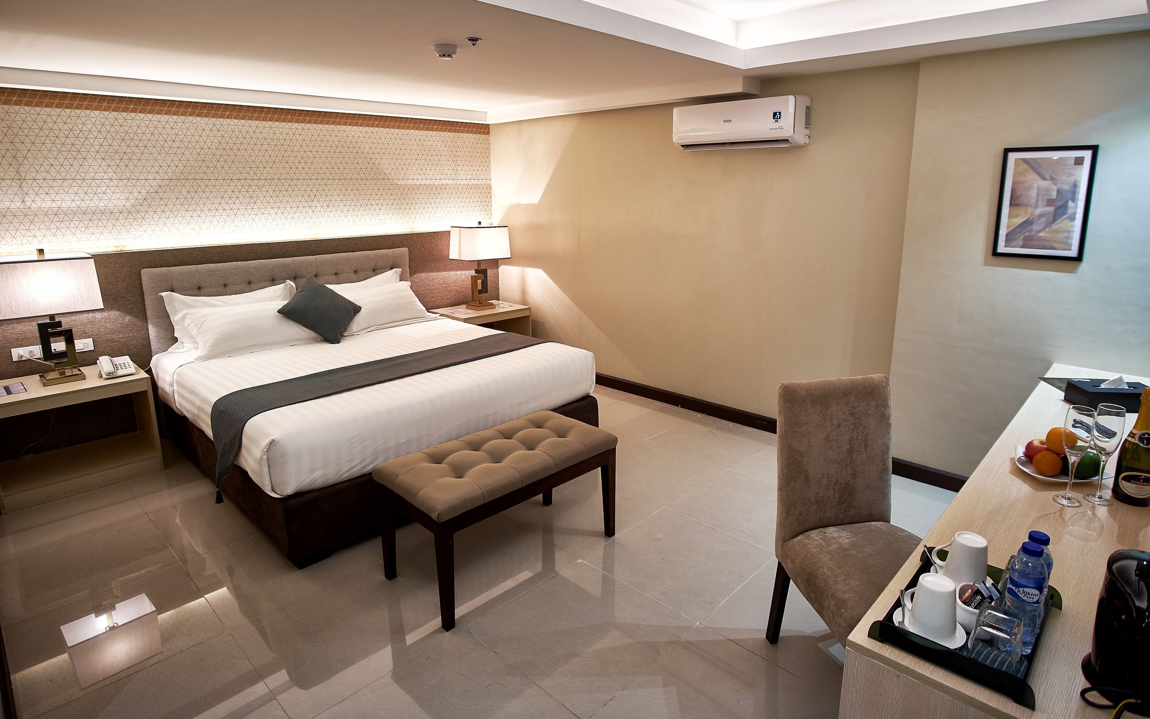 A hotel room with a large bed, a bedside table with a lamp, a chair, and a desk with a tray of water bottles and glasses.