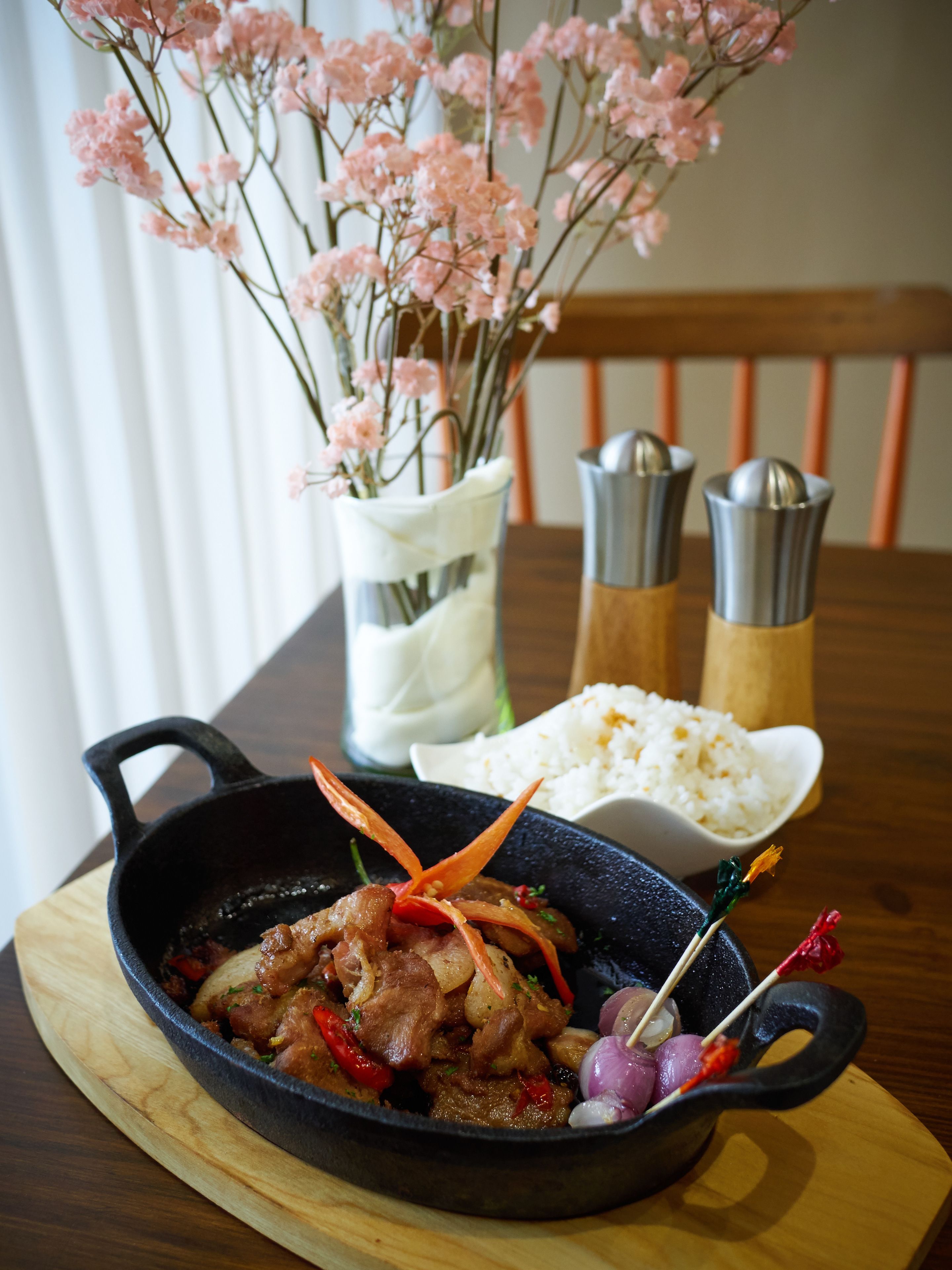 A meal with meat and vegetables in a cast iron skillet, a bowl of rice, and a vase with pink flowers.