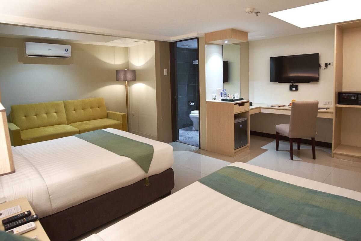 A hotel room with two beds, a sofa, a desk, a chair, a TV, and an air conditioning unit.