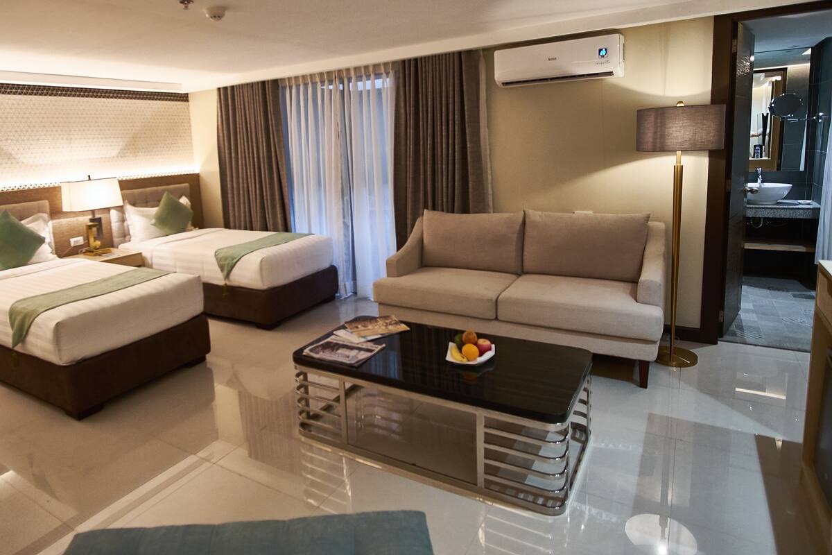 A hotel room with two beds, a sofa, a coffee table, and a bathroom visible through an open door.