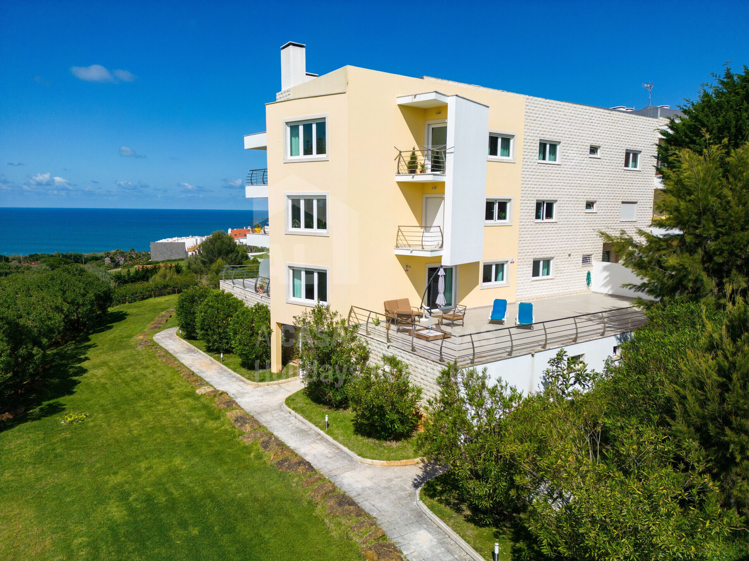 A modern multi-story building with balconies, a patio area with outdoor furniture, and a view of the sea.