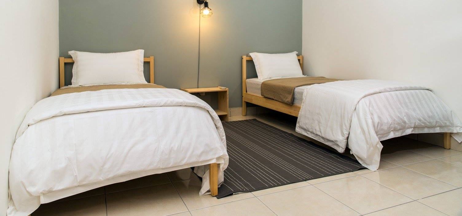 A room with two single beds, each with a wooden frame and white bedding. There is a small bedside table with a lamp on one side and a shelf on the other. The room has a tiled floor and a dark striped rug in the center.