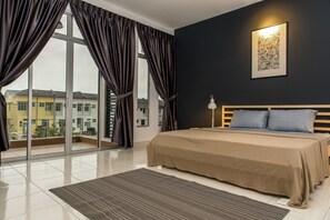 A bedroom with a large window, a bed with a beige bedspread, and a nightstand with a lamp.