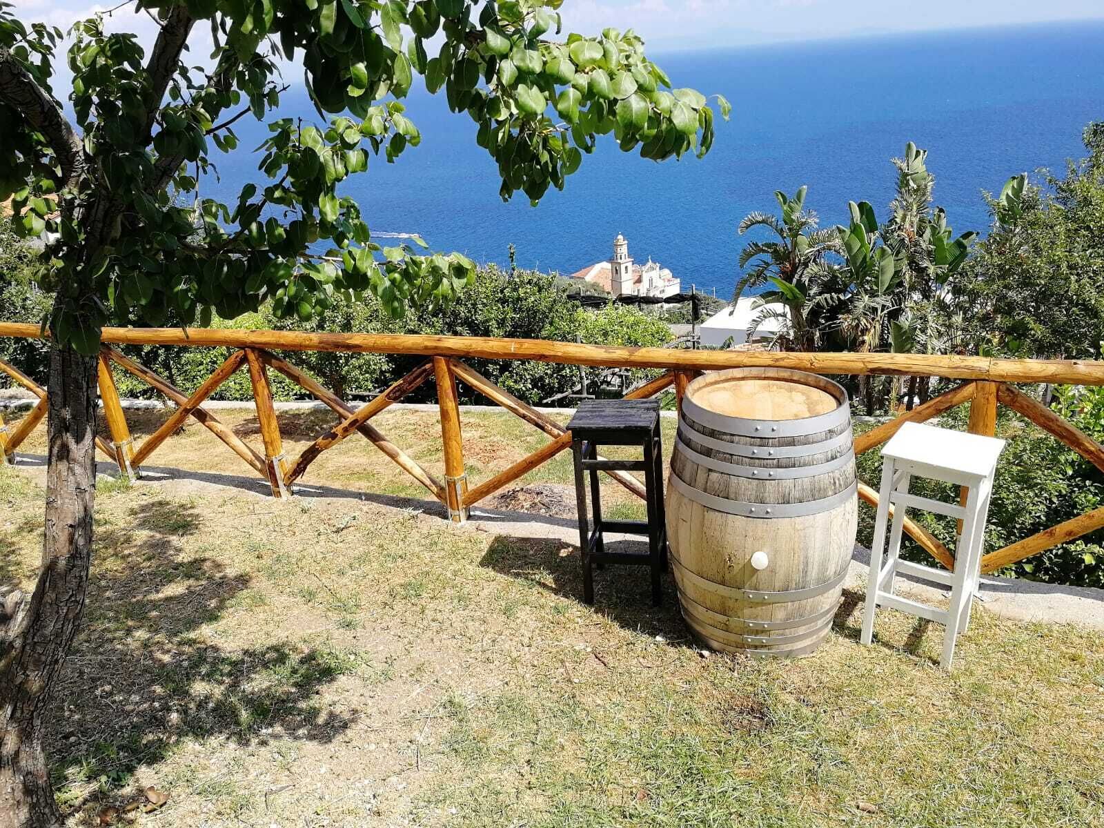 A rustic outdoor seating area with a wooden fence, a wooden barrel, and a small stool, overlooking a coastal landscape with a church and ocean view.