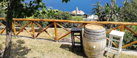 A rustic outdoor seating area with a wooden fence, a wooden barrel, and a small stool, overlooking a coastal landscape with a church and ocean view.