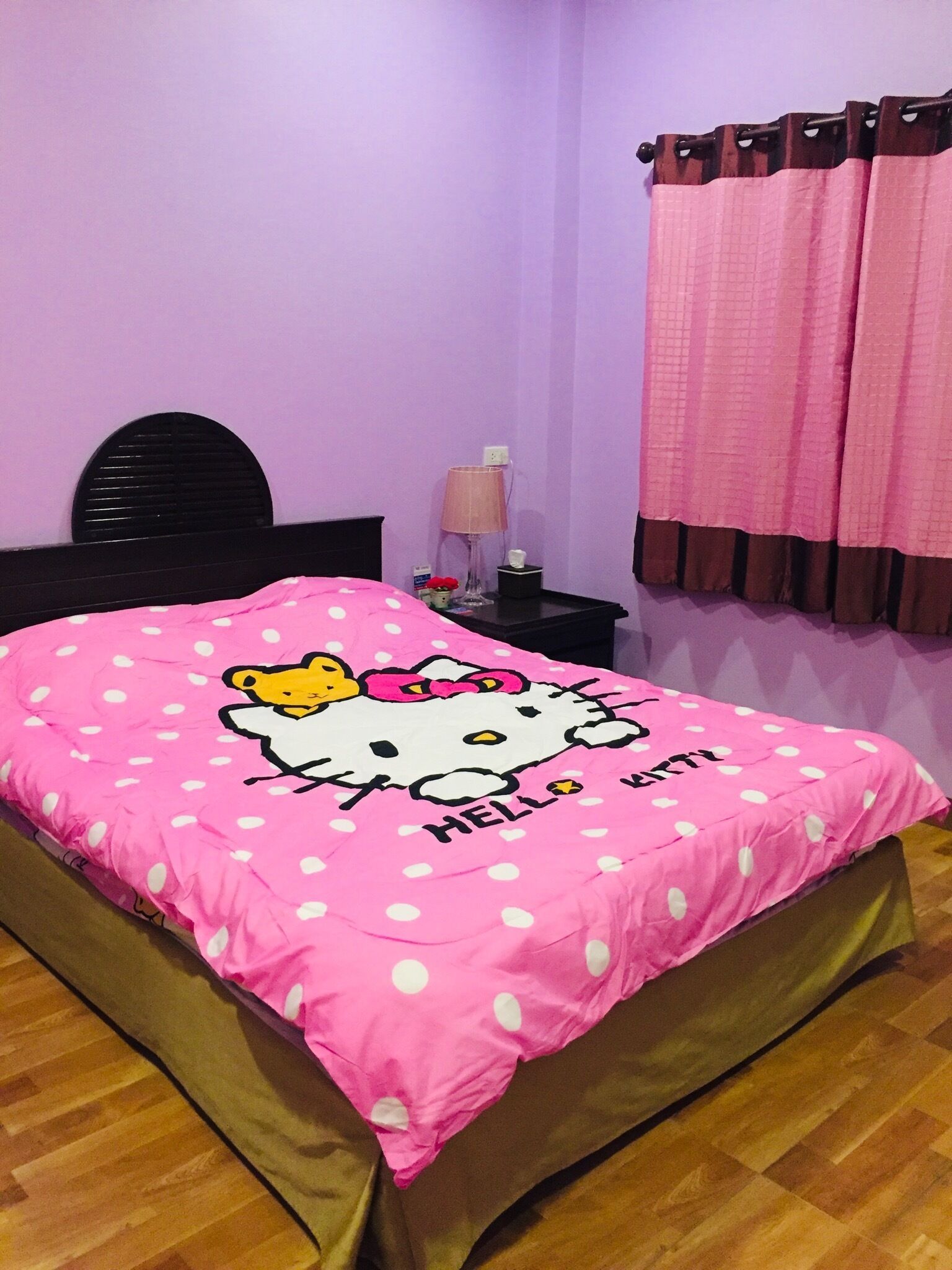 A bedroom with a bed covered in a Hello Kitty-themed pink blanket, a black headboard, a bedside table with a lamp, and a window with pink curtains.