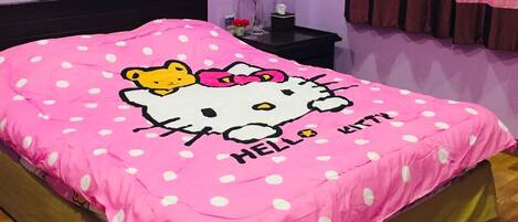 A bedroom with a bed covered in a Hello Kitty-themed pink blanket, a black headboard, a bedside table with a lamp, and a window with pink curtains.