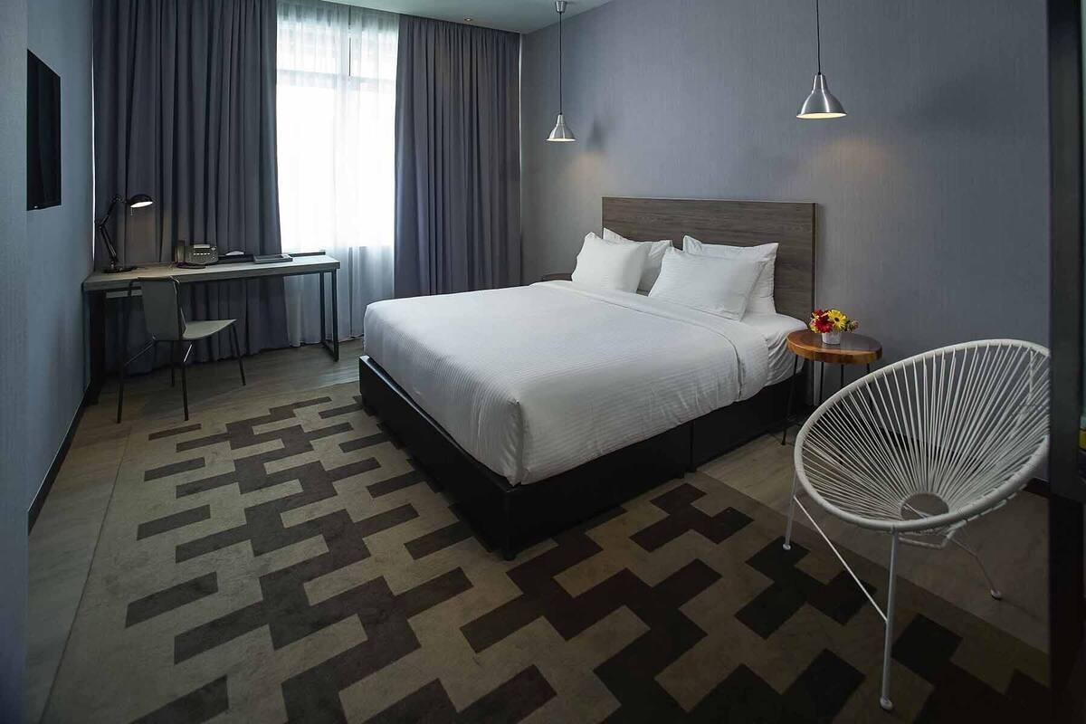 A modern hotel room with a large bed, a desk, a chair, and a small table with flowers.