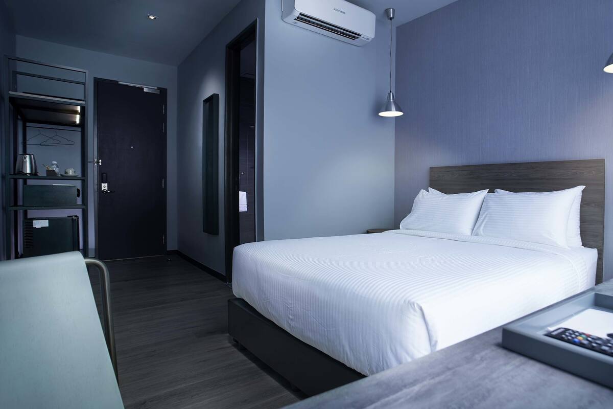 A modern hotel room with a large bed, bedside table with a remote control, a small wooden side table with a potted plant, and a desk with a chair.
