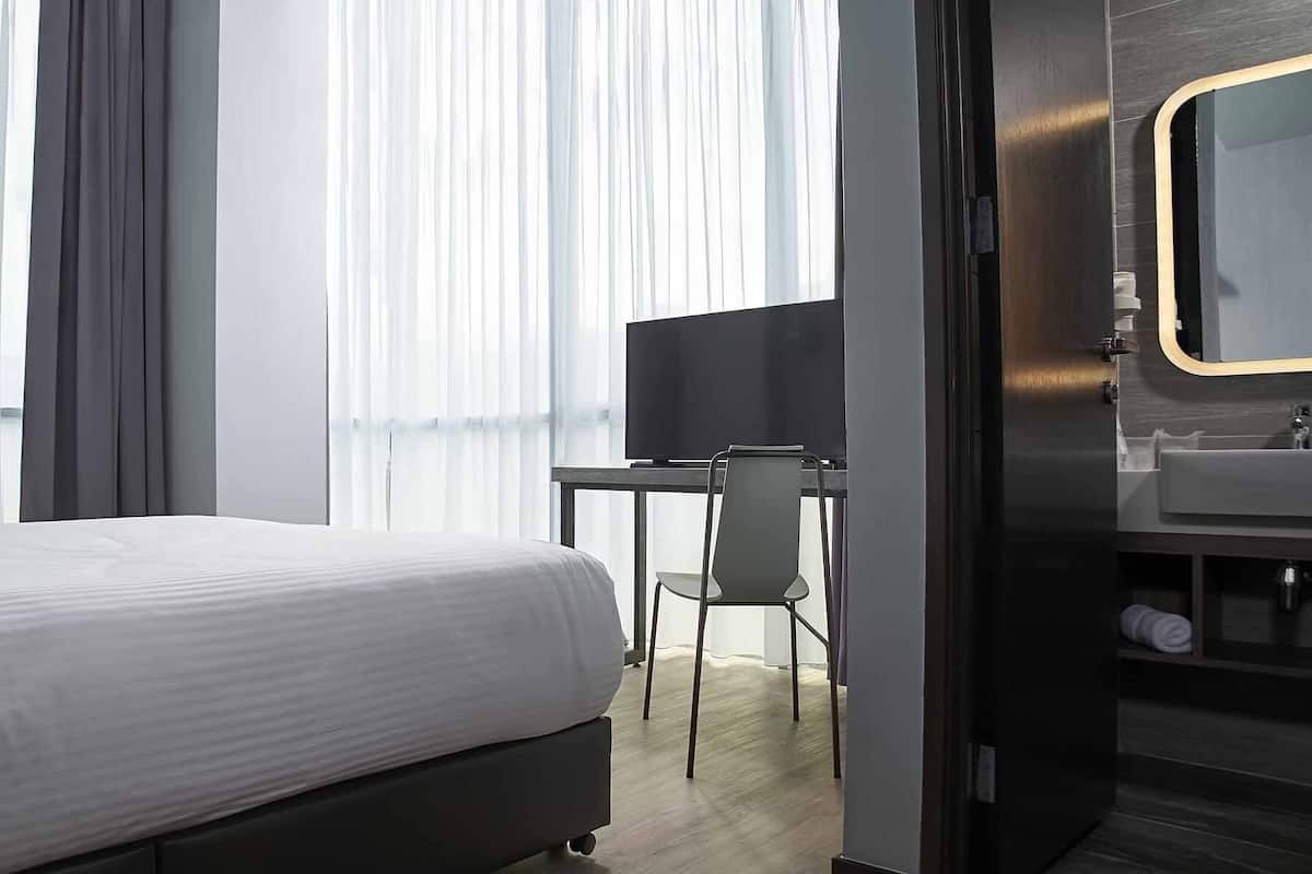 A modern hotel room with a bed, a desk, a chair, a TV, and a bathroom with a mirror and sink.
