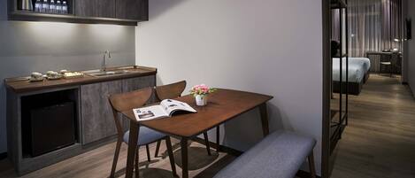 A modern hotel room with a wooden dining table, two chairs, a bench, a small kitchen area with a sink and a coffee machine, and a bedside table with a lamp.