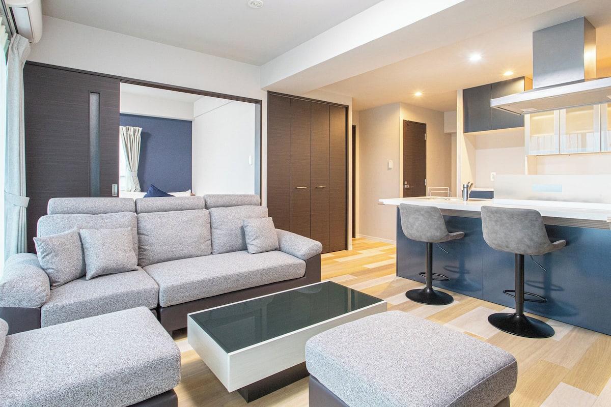 Een moderne woonkamer met een grijze bank, een glazen salontafel, en een keukenhoek met een bar en krukken.