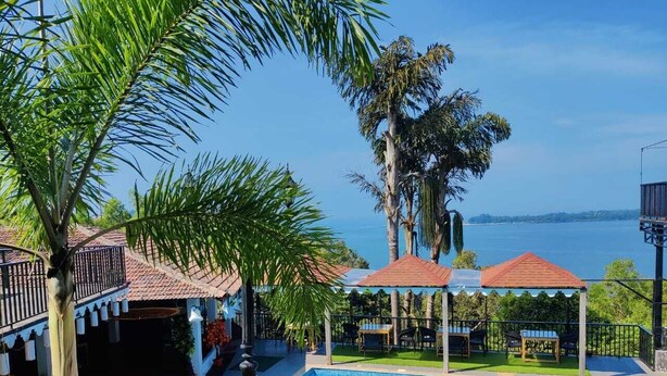 A pool area with a view of the sea, surrounded by palm trees and covered seating areas. - Raposo Holiday Resort (Vagator)