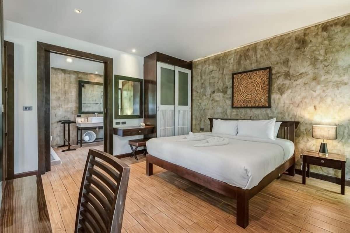 Un dormitorio con una cama grande, un escritorio, una silla, un espejo y un baño visible a través de una puerta abierta.