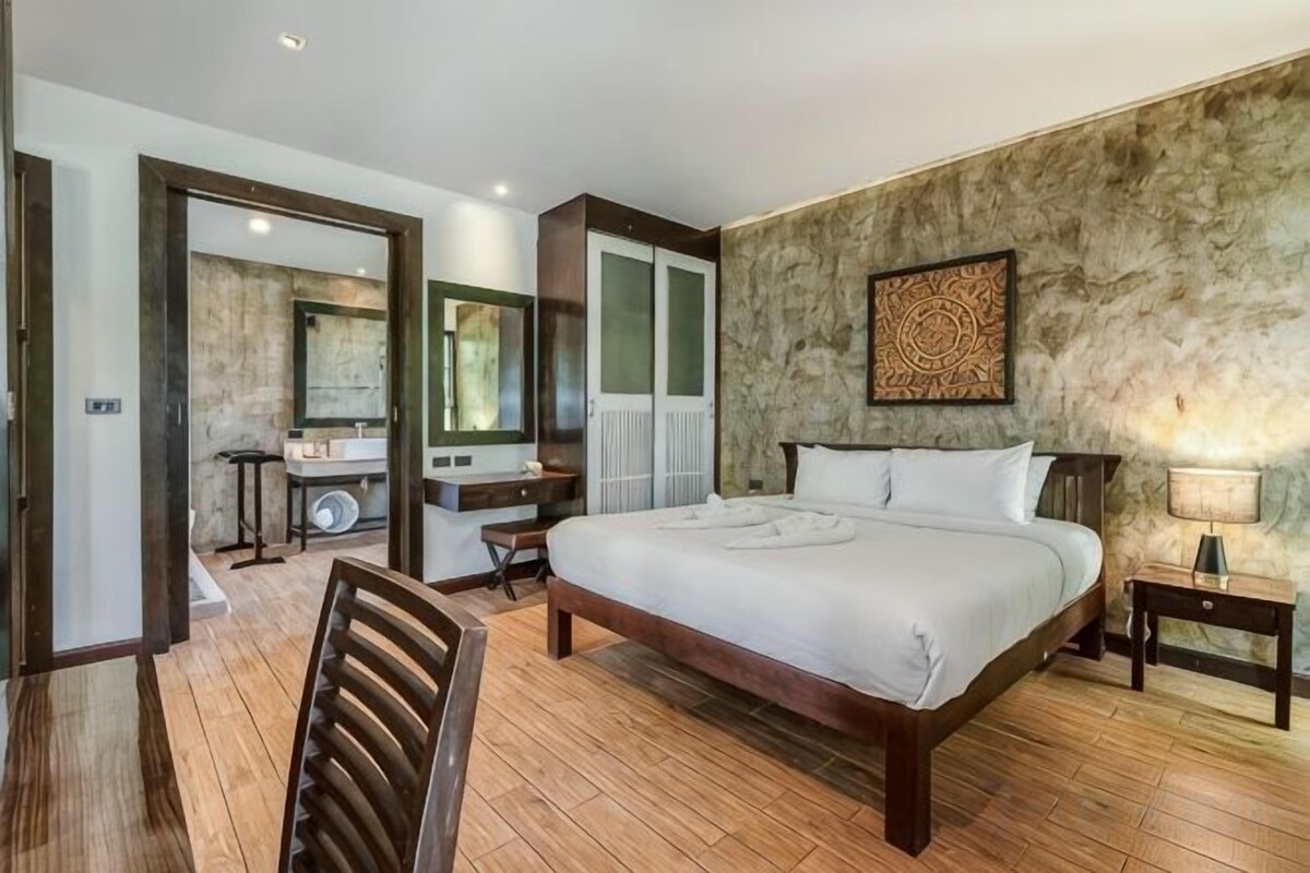 Un dormitorio con una cama grande, un escritorio, una silla, un espejo y un baño visible a través de una puerta abierta.