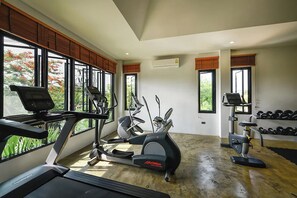 A gym with exercise equipment, large windows, and a view of trees. - Coco Retreat Phuket Resort and Spa Sha+ (Chalong)