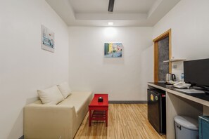A compact hotel room with a sofa, a small red table, a kitchenette with a fridge, a TV, and artwork on the walls. - Coco Retreat Phuket Resort and Spa Sha+ (Chalong)
