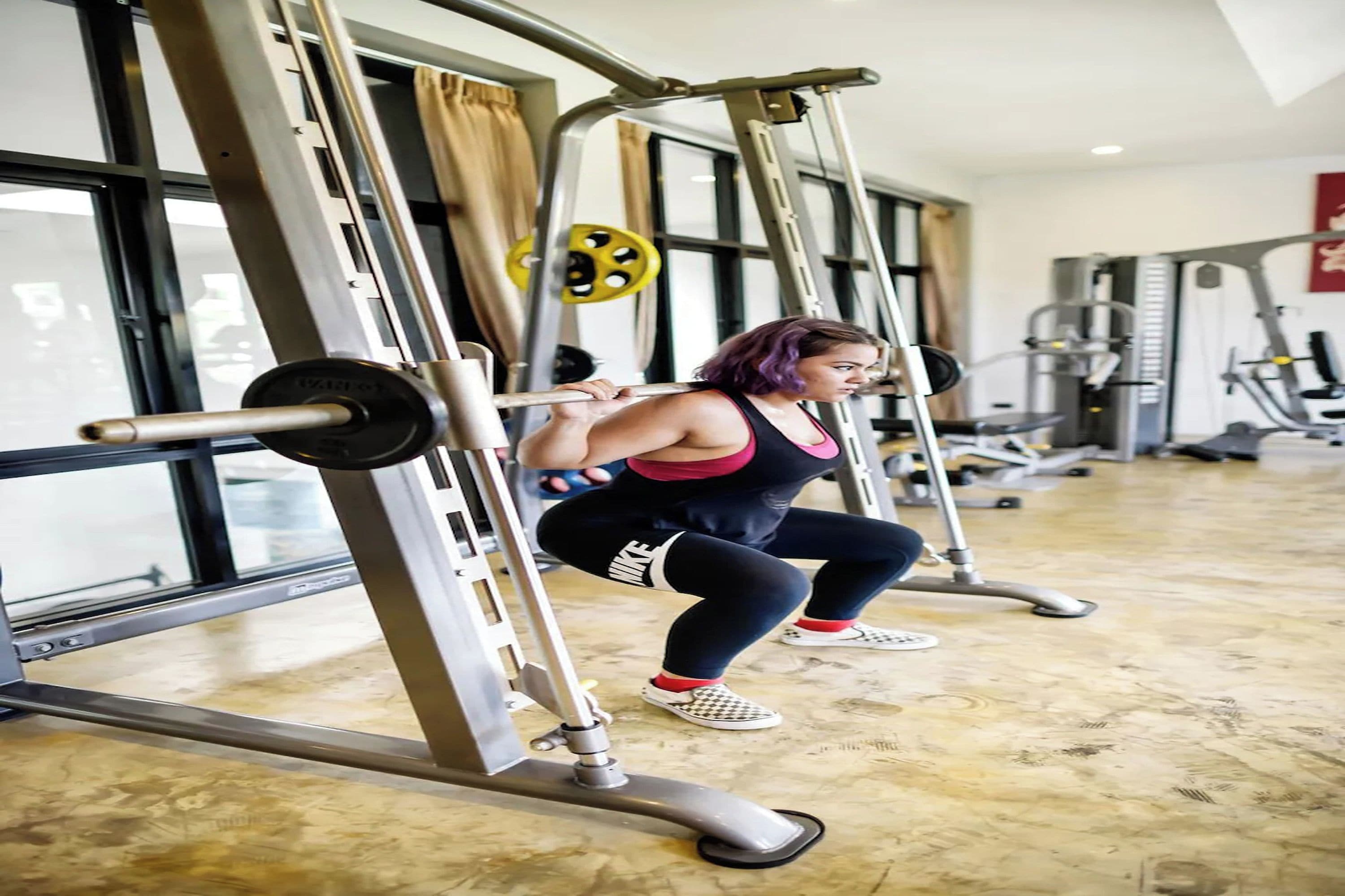 A person exercising on a squat machine in a gym with large windows and various exercise equipment.