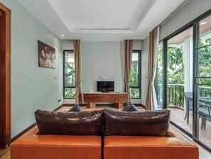 A modern living room with a sofa, a TV on a wooden cabinet, and large windows overlooking a balcony with greenery. - Coco Retreat Phuket Resort and Spa Sha+ (Chalong)