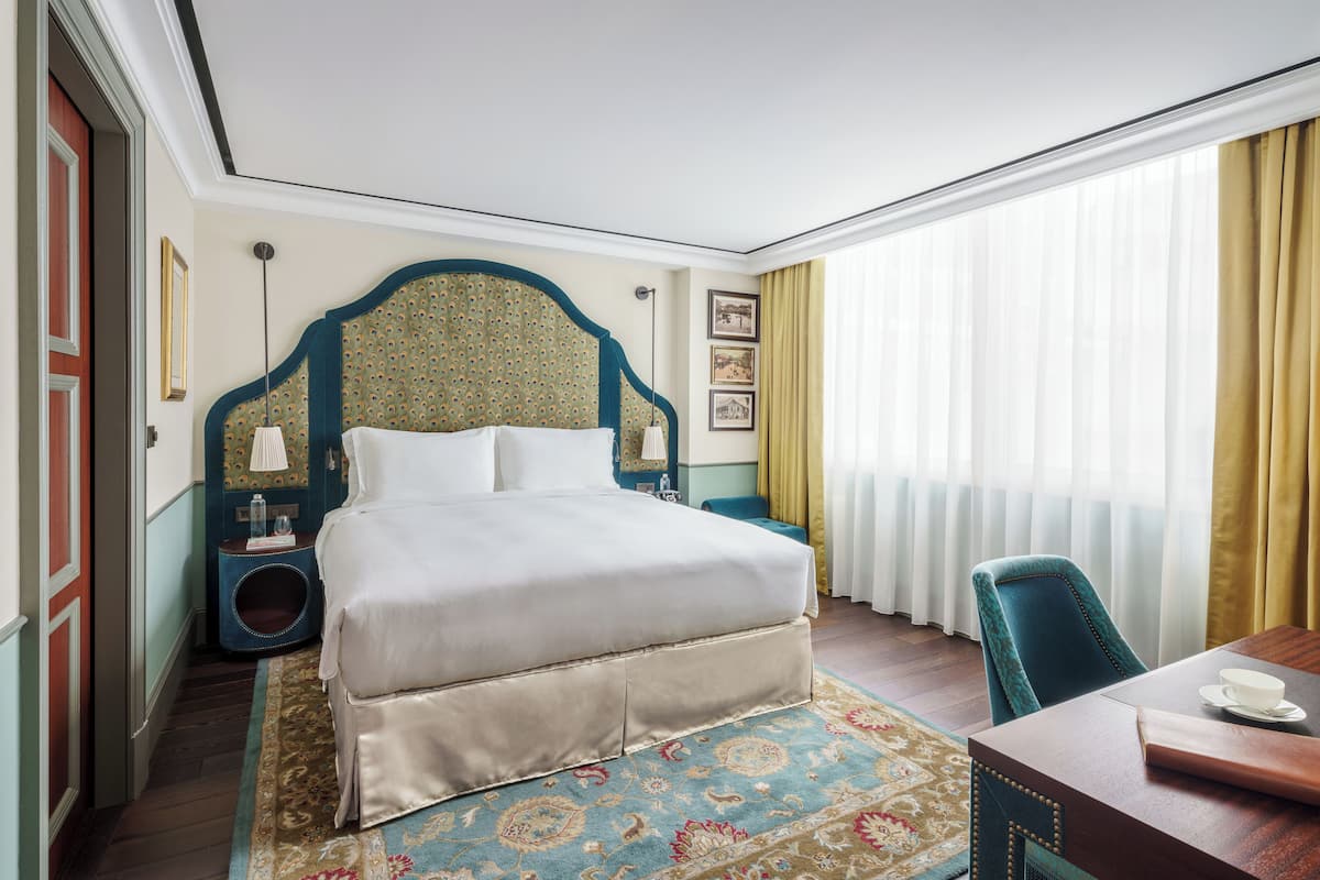 Ein Hotelzimmer mit einem großen Bett, einem Schreibtisch mit Stuhl, einem kleinen Nachttisch, einer Lampe und einem Fenster mit Vorhängen.