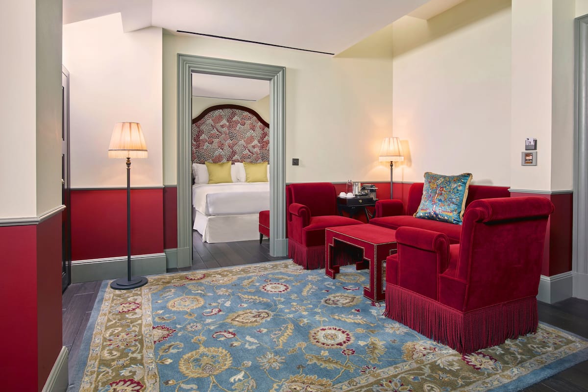 Ein Hotelzimmer mit rotem Sofa, einem kleinen Tisch, einem dekorativen Teppich und einem Bett mit gemustertem Kopfteil.
