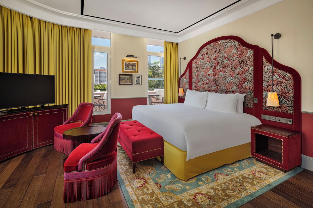 Ein Hotelzimmer mit einem großen Bett, einem Fernseher, einem rot gepolsterten Sessel, einem kleinen runden Tisch und Blick auf die Stadt durch große Fenster.